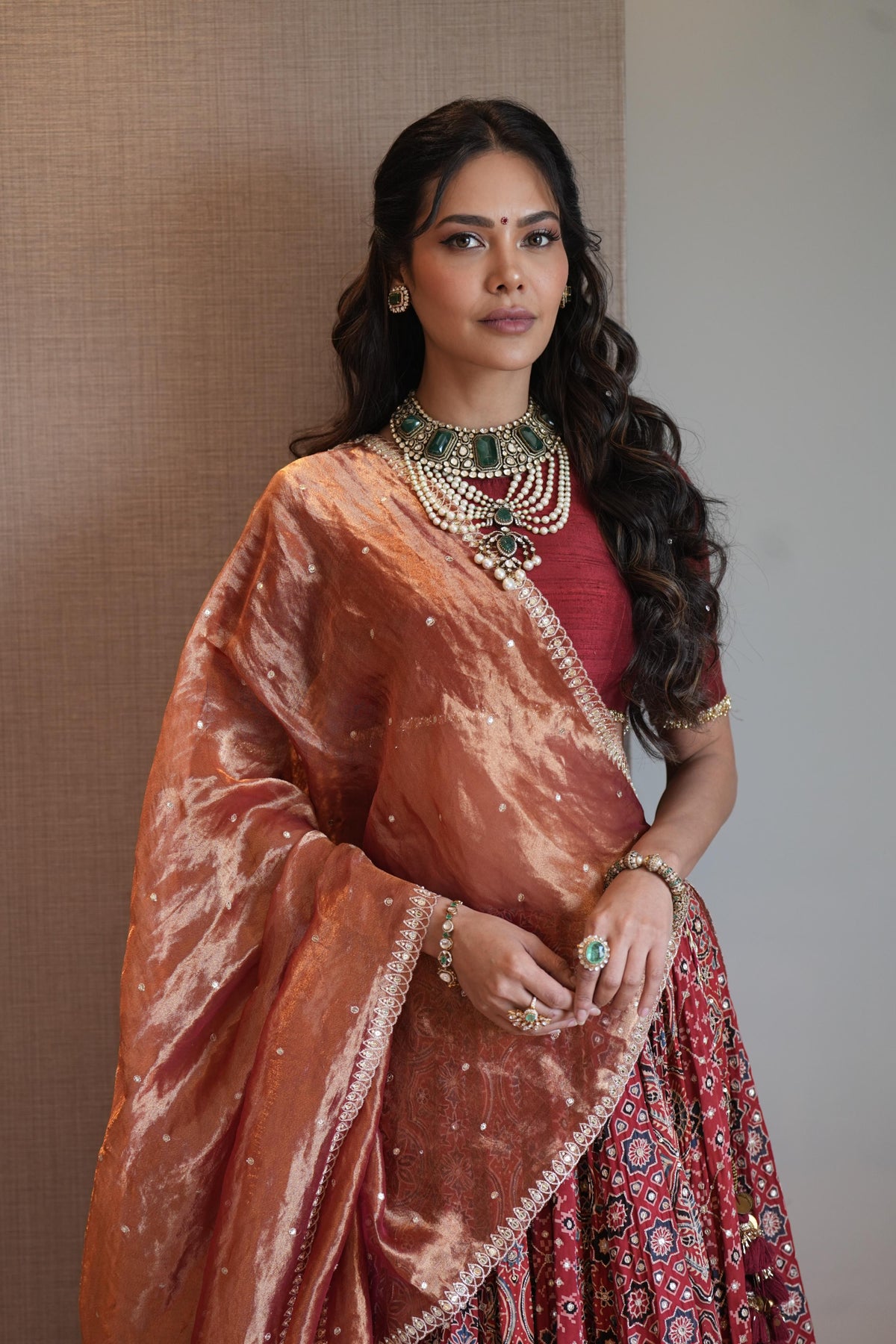 A woman modeling a traditional Indian outfit featuring a rust-orange marbled dupatta, a red embroidered blouse, and a red patterned lehenga skirt, accessorized with a statement green and gold necklace.