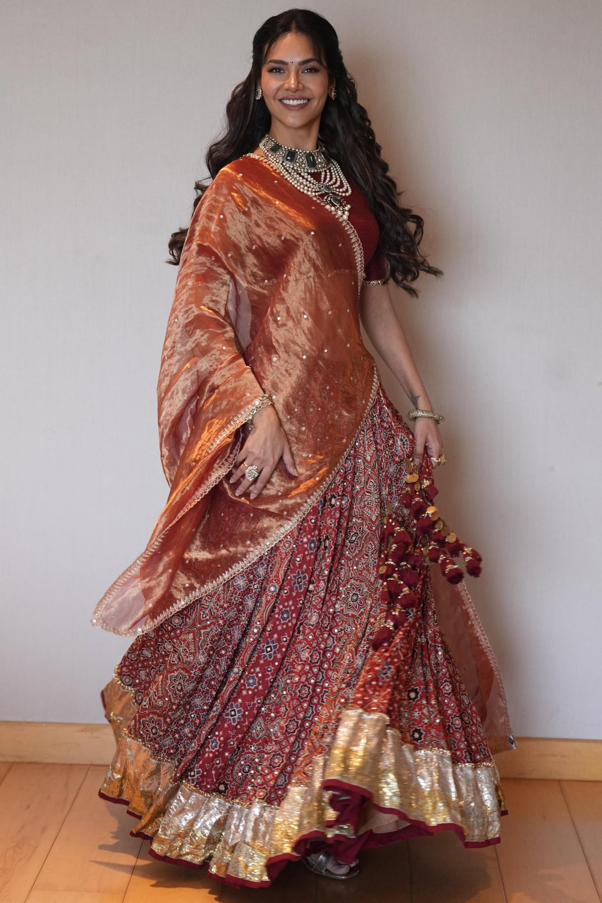 A woman modeling a traditional Indian lehenga choli set in a studio setting. The outfit features a metallic copper-orange embroidered choli with a matching dupatta and a heavily embellished red and gold lehenga skirt.
