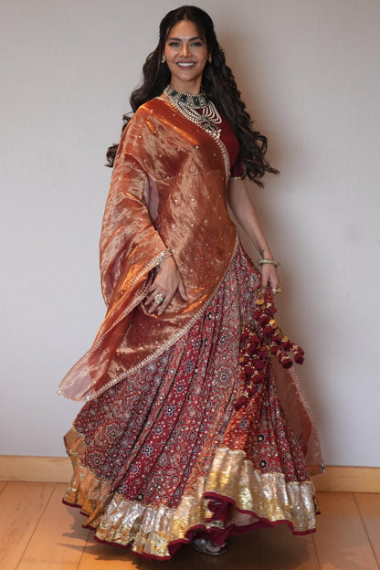 A woman modeling a traditional Indian lehenga choli set in a studio setting. The outfit features a metallic copper-orange embroidered choli with a matching dupatta and a heavily embellished red and gold lehenga skirt.