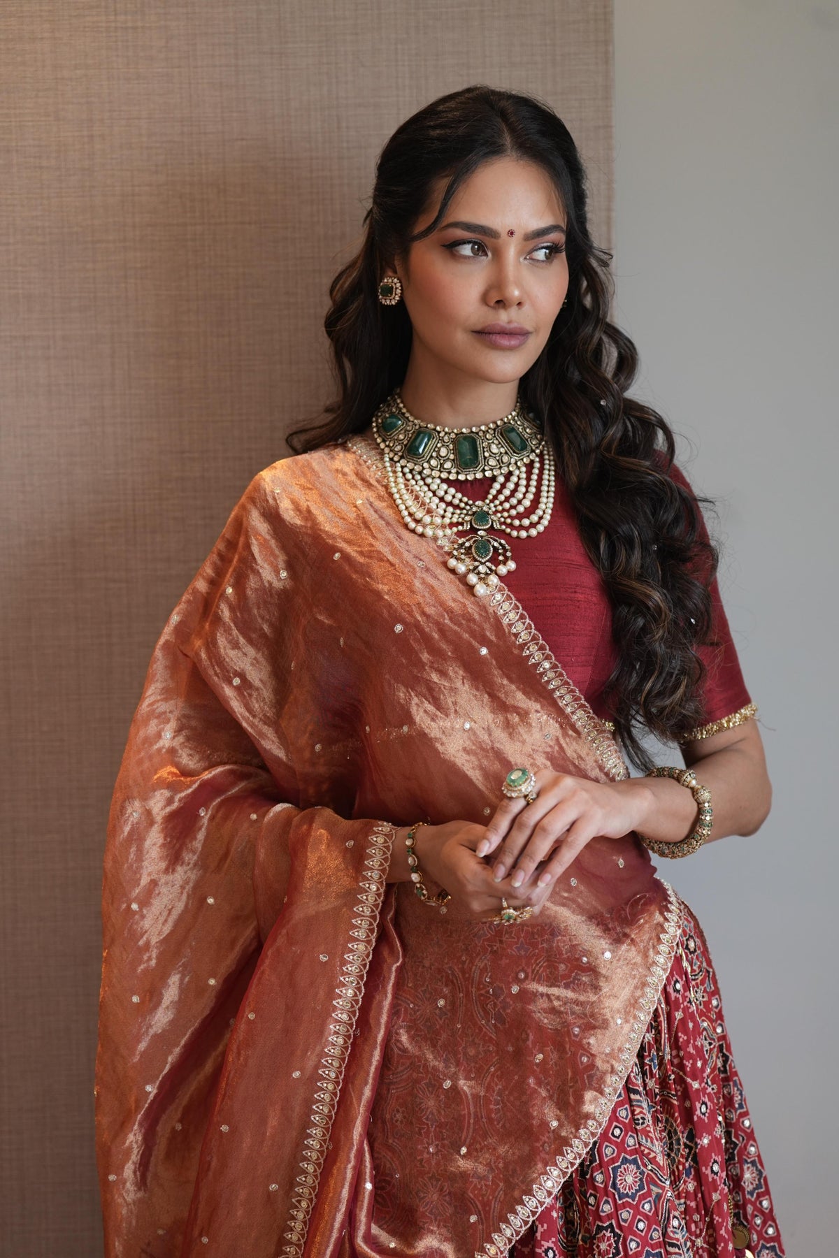 A woman wearing traditional Indian attire, featuring a metallic bronze dupatta draped over her shoulders, a deep red blouse, and a red patterned skirt, accessorized with a green and gold statement necklace and gold jewelry.