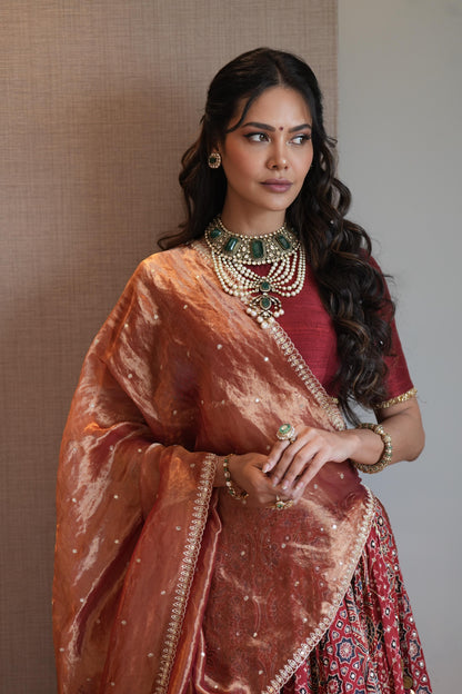 A woman wearing traditional Indian attire, featuring a metallic bronze dupatta draped over her shoulders, a deep red blouse, and a red patterned skirt, accessorized with a green and gold statement necklace and gold jewelry.
