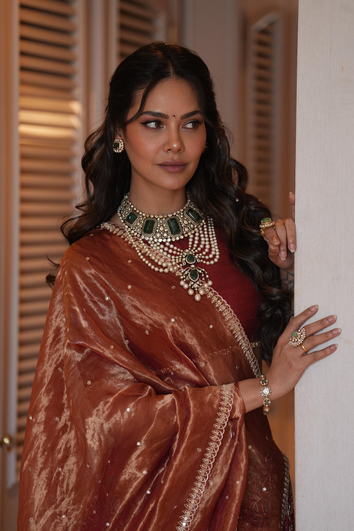 A woman wearing an ornate bronze-brown embellished sari with a red inner garment and layered gold and green jewelry, posing indoors against a light-colored wall.